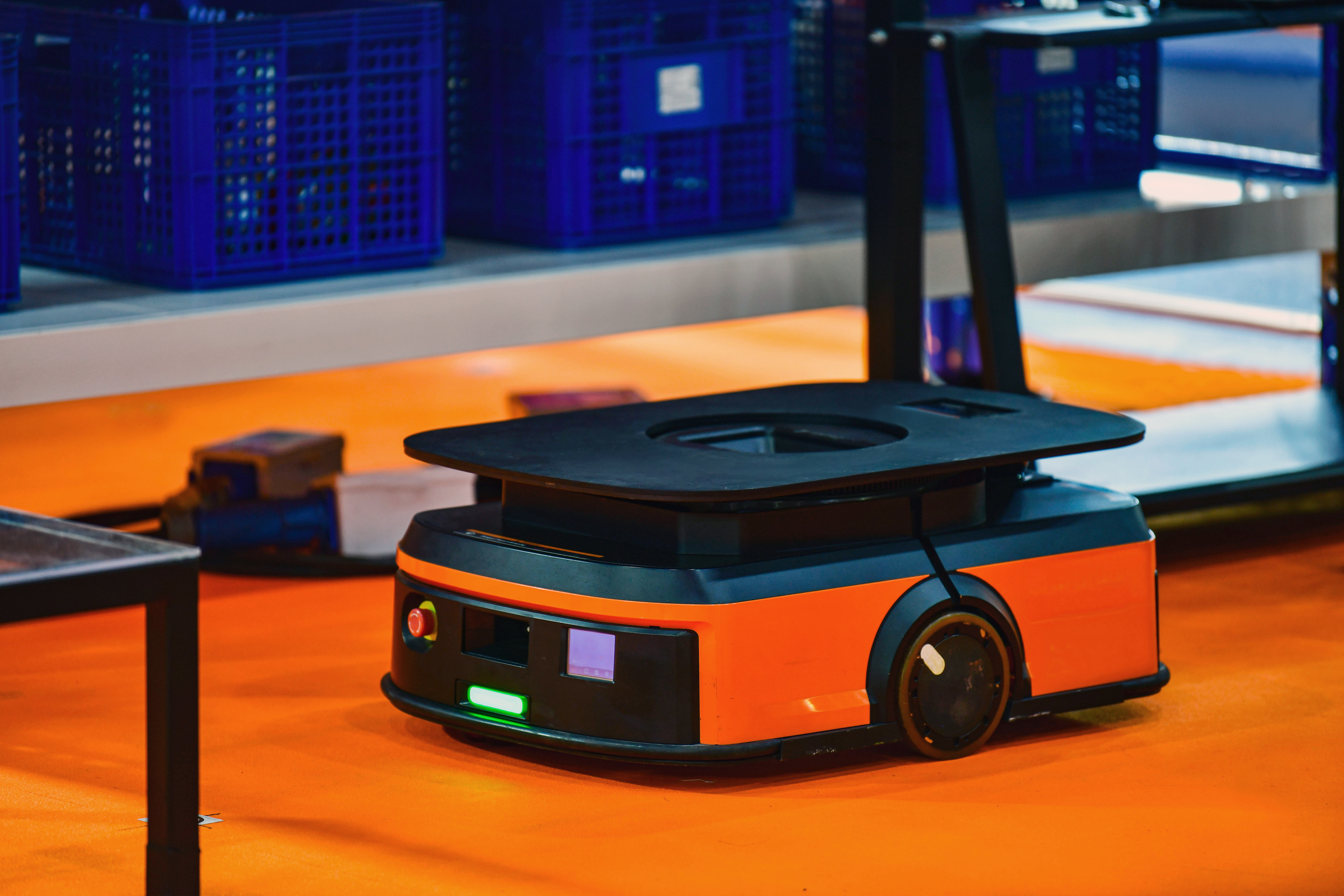 Autonomer mobiler Transportroboter in Orange und Schwarz auf dem Boden einer Industriehalle mit Lagerbehältern im Hintergrund.