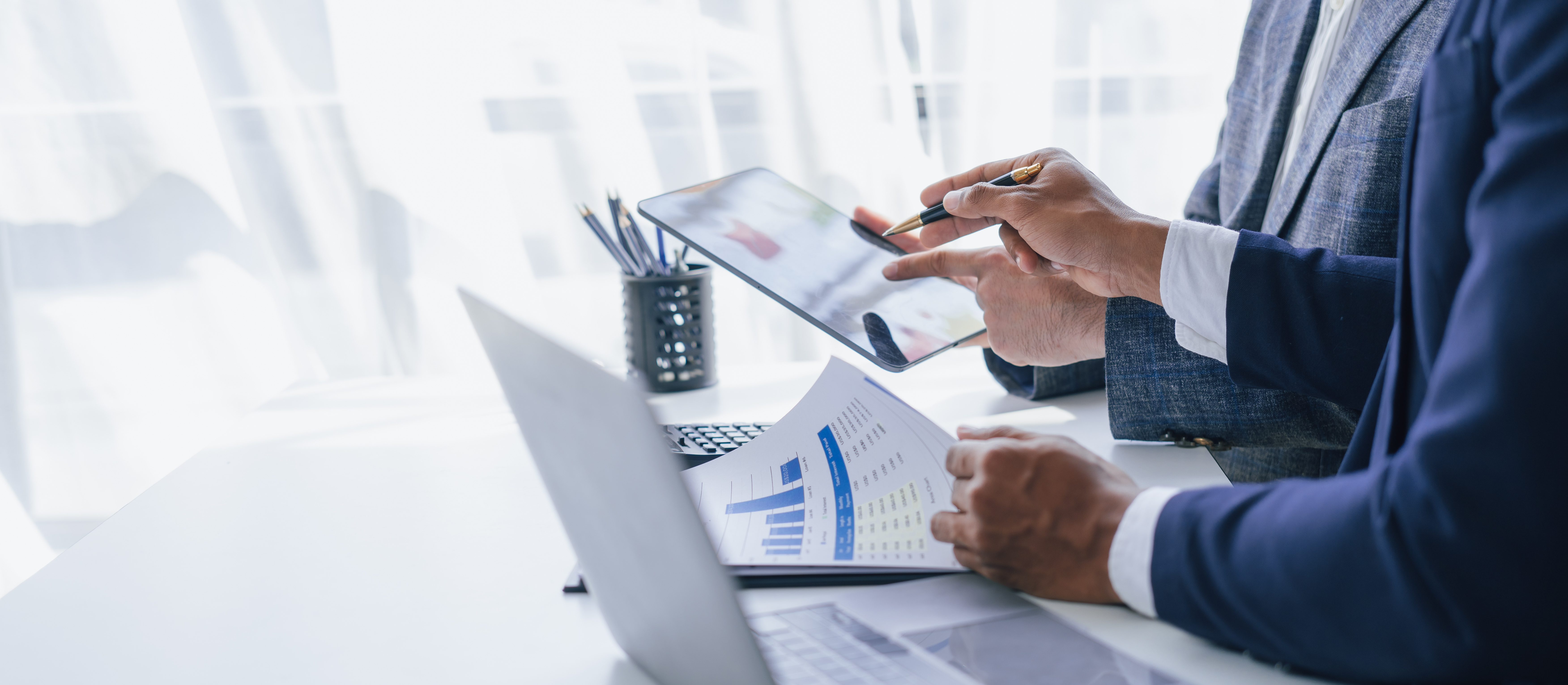 Zwei Geschäftsleute analysieren Finanzberichte und Diagramme an einem Schreibtisch mit Laptop und Tablet.