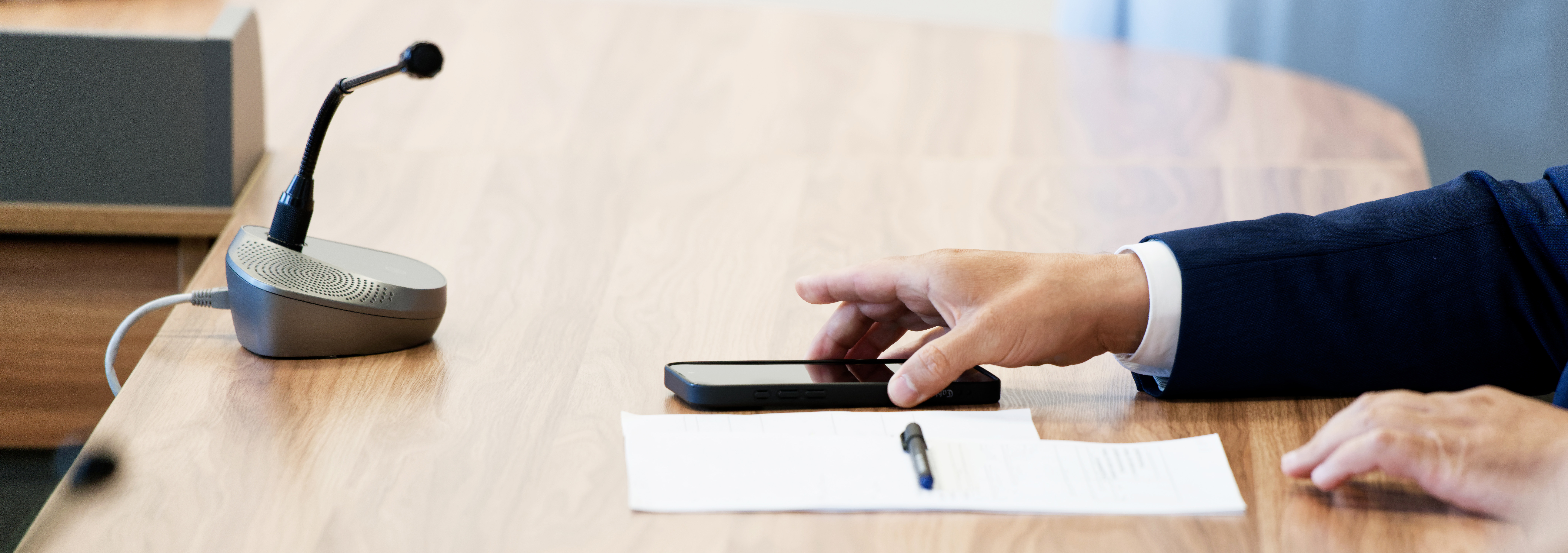 Geschäftsmann im Anzug bedient ein Smartphone an einem Konferenztisch mit Mikrofon, Dokumenten und Stift.