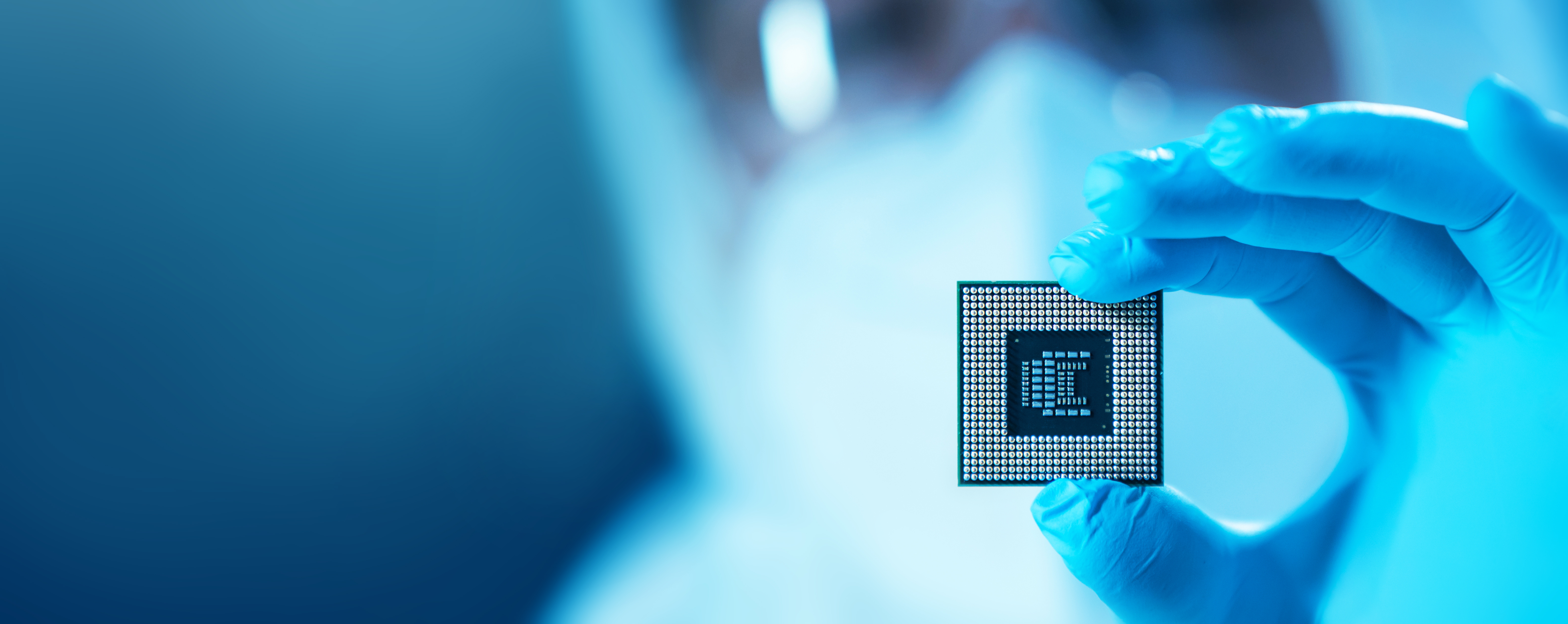 Nahaufnahme einer Hand mit blauem Schutzhandschuh, die einen Mikrochip zwischen zwei Fingern hält.