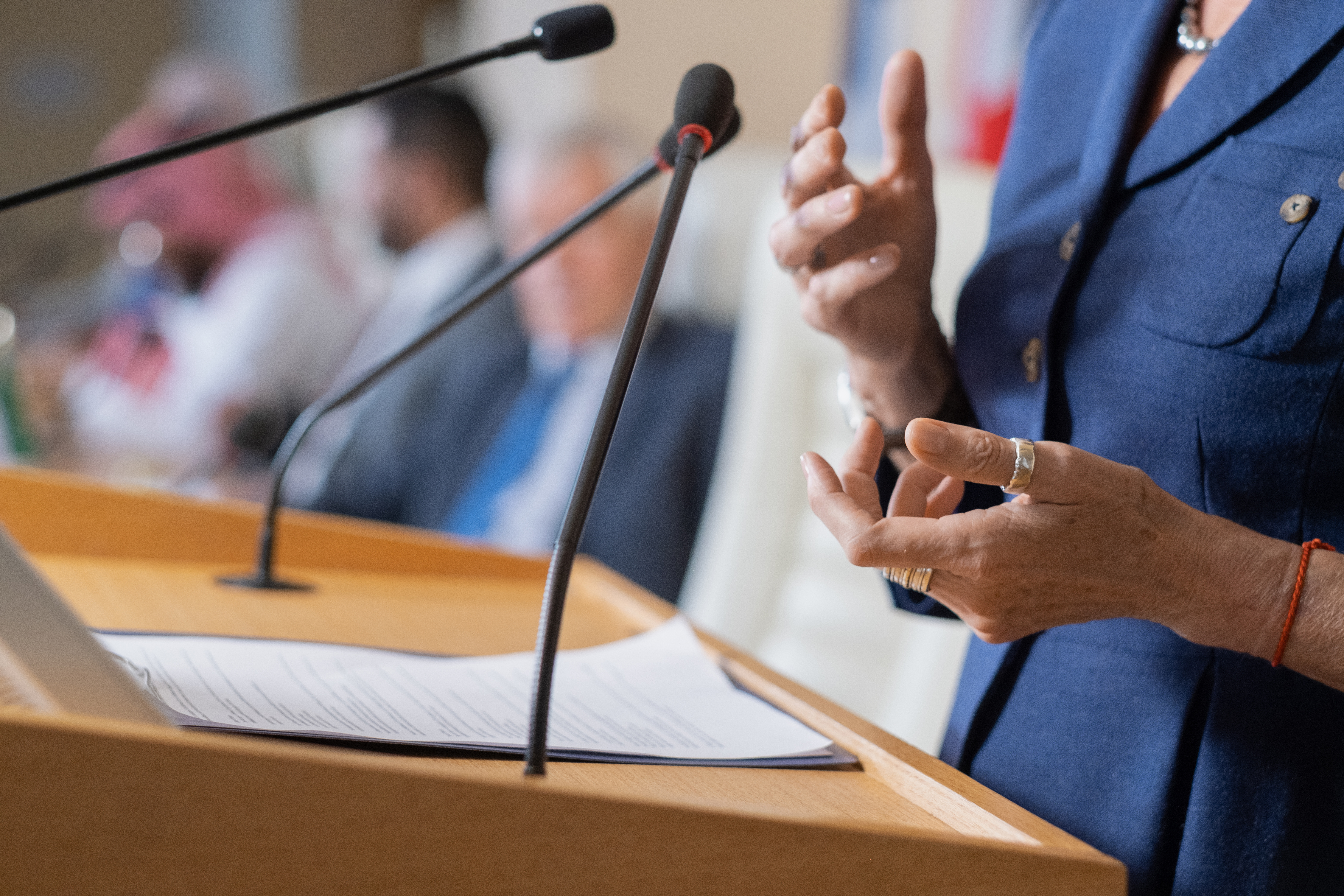 Eine Person im blauen Blazer hält eine Rede an einem Rednerpult mit Mikrofonen und gestikuliert vor einem Publikum.