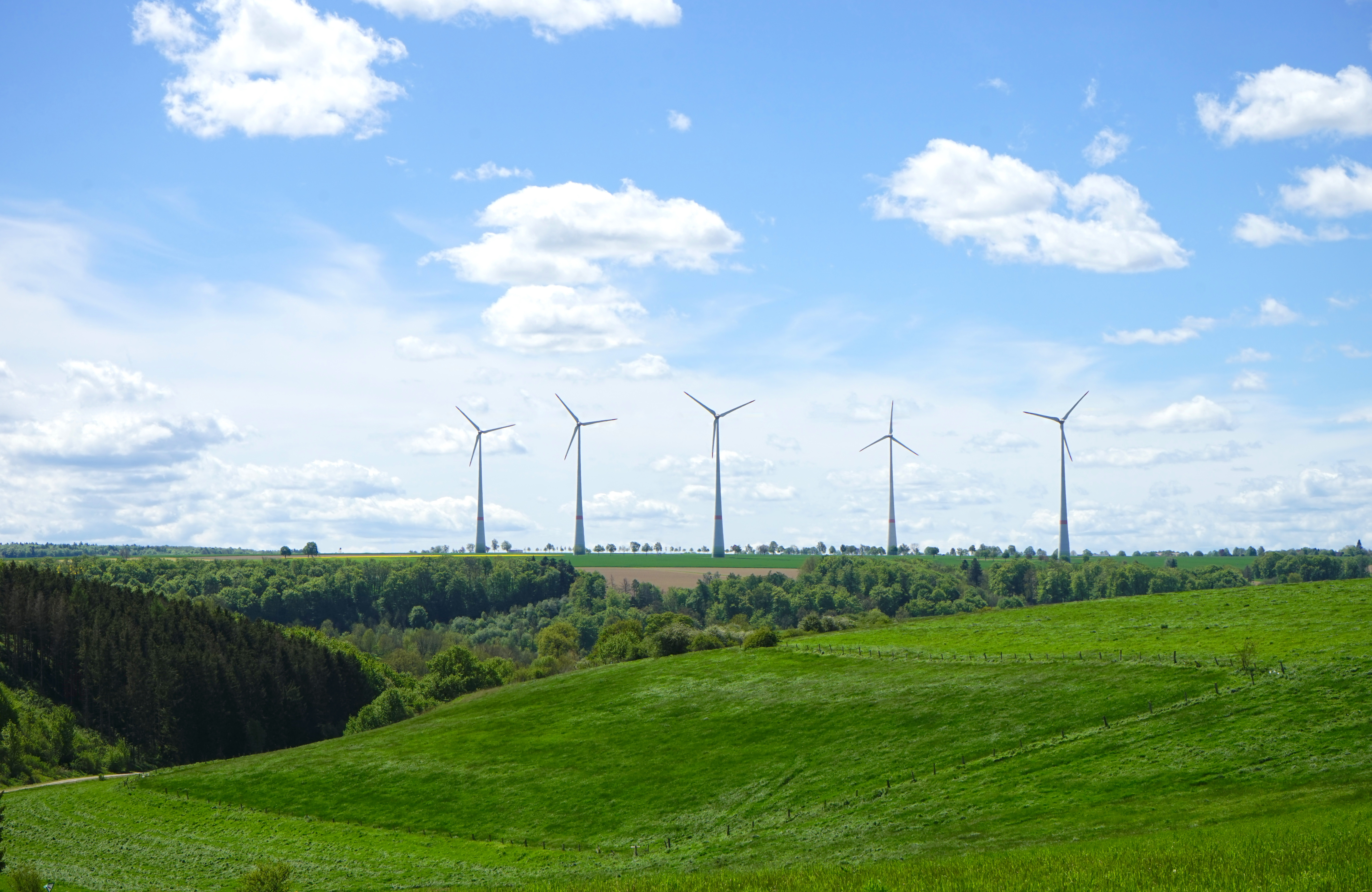 Fünf Windkraftanlagen stehen auf einer Anhöhe zwischen grünen Wiesen und Wäldern unter einem blauen Himmel mit Wolken.