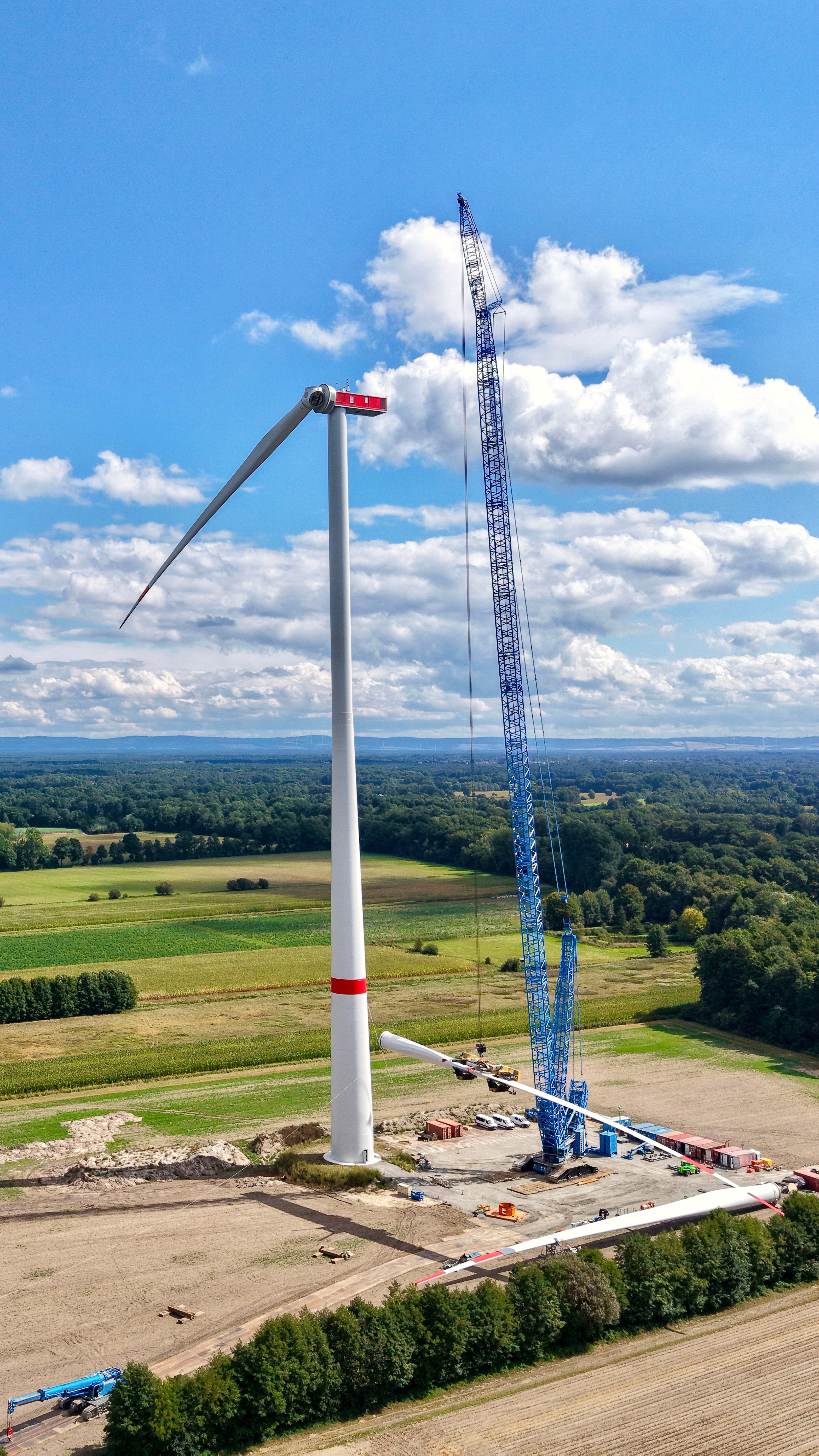 Luftaufnahme einer Windkraftanlage im Bau mit großem Montagekran und mehreren Rotorblättern auf einer Baustelle zwischen Feldern und Wäldern.